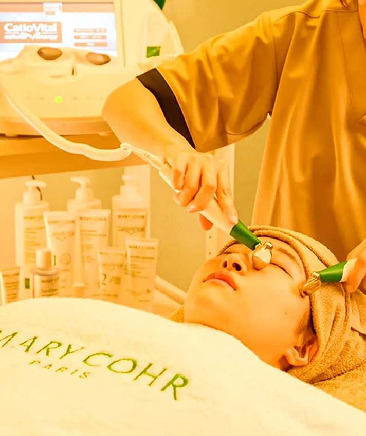 エステブランド「MARY COHR」の施術用タオルに包まれ、目元にローラー器具でフェイシャルケアを受ける女性