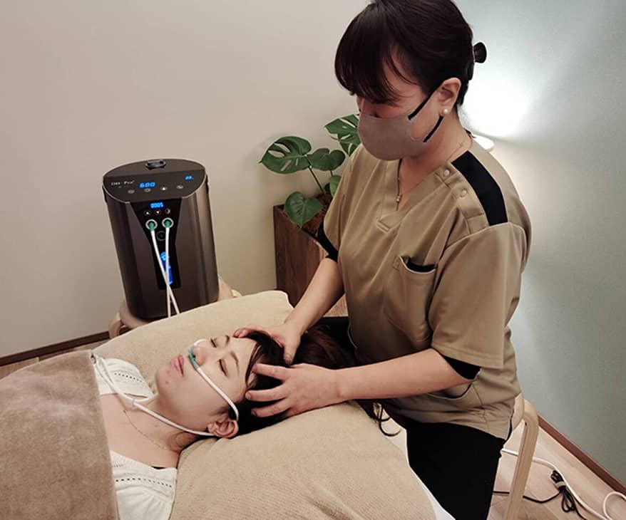 酸素吸入器を使用しながら、エステティシャンが女性にヘッドマッサージを行っている施術風景