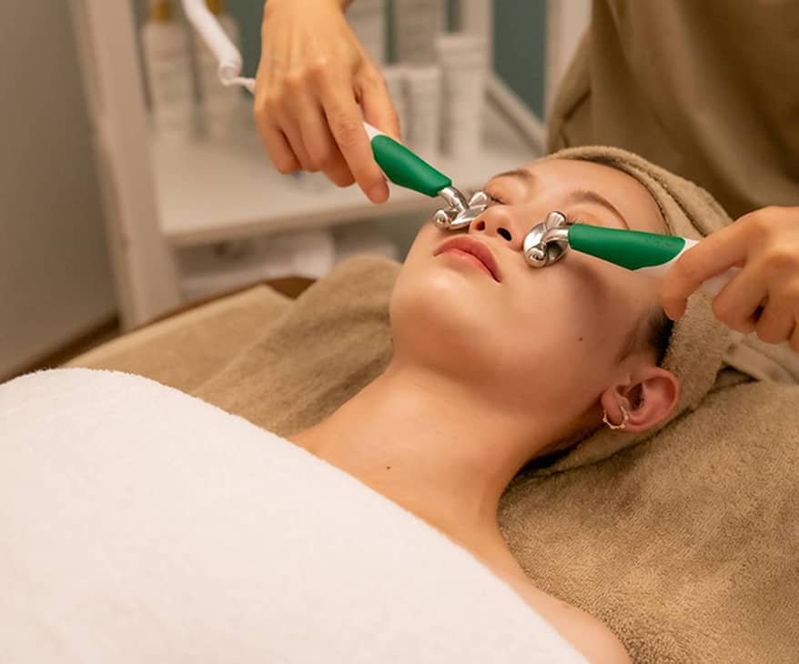 リラックスした表情でフェイシャルマッサージを受ける女性と、緑色のハンドルローラーを使う施術者